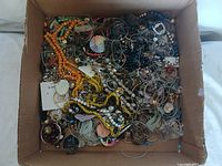 Photo showing assorted jewelry including beaded necklaces, hoop earrings, and mixed pendants piled inside a cardboard box.