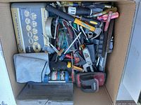Top-down photo of assorted hand tools in cardboard box showing many small tools including screwdrivers, pliers, hammers, and measuring tape.