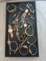 Top view of assorted women's wristwatches in a black tray showing variety of bracelet styles and watch faces.