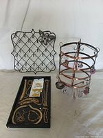 Photo showing black metal mesh jewelry display grid with a floral decoration, flat black tray containing assorted chains, bracelets, and a carded set of jewelry.