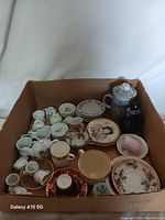 Top view of the assortment featuring varied tea cups, saucers, creamers, a teapot, and small decorative plates inside a cardboard box.