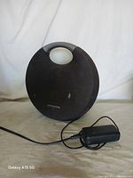 Full front view of Harman/Kardon portable speaker with power adapter connected and visible.