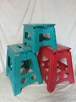Group photo of three plastic folding stools, two teal and one red, stacked together showing their size and condition with visible paint marks.