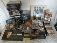 Photo showing assorted media including DVD and Blu-Ray cases stacked, cassette tapes arranged vertically, and multiple box sets laid out including Garth Brooks, Hank Williams, Led Zeppelin, and The Who.