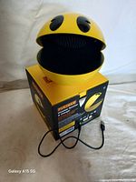 Pac-Man wireless speaker in yellow, open mouth showing speaker grill with attached charging cable, sitting on original box.