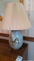 Full view of ceramic pottery lamp with beige pleated shade on wooden surface.