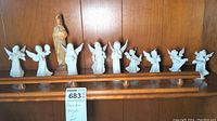 Ten porcelain figurines displayed on a wooden shelf; nine Dresden angels with wings, mostly playing musical instruments, and one religious figure holding a child.
