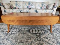 Front view of medium brown wooden drop leaf coffee table in front of floral sofa, showing table top and tapered legs with slight wear.