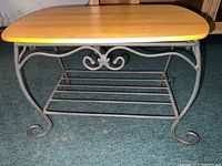 Side table with wooden top and curved metal base with decorative scroll details on the sides, sitting on a green carpet.