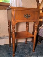 Front and angled view of colonial style nightstand showing one drawer with metal pull and lower shelf, maple tone wooden finish, turned legs.
