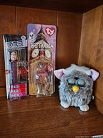 Photo showing Furby toy, Star Wars PEZ candy dispenser with candy, and Britannia the mini Beanie Baby in original packaging all together on a wooden shelf.