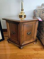 Side view showing hexagonal shape, paneled wooden sides, metal handles, and bun feet on hardwood floor next to floral couch and brass lamp on top.