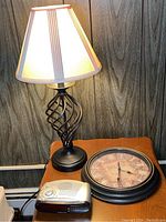 Photo of metal table lamp lit, showing twisted cage base and beige lampshade with vertical stripes, along with Emerson electronic clock radio and round decorative wall clock on a table