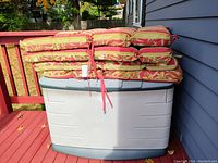 Photo of a large plastic outdoor storage box with four stacked reversible patio chair cushions on top. Cushions are red and yellow patterned, padded, with ties. Storage box lid is closed but visibly damaged.