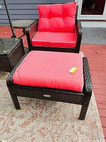 Dark wicker framed armchair with tufted red back and seat cushions with white piping, positioned on an outdoor deck with a patterned rug underneath, accompanied by a matching ottoman with red cushion of similar design.
