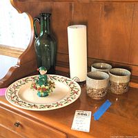 Photo showing entire lot including holly serving plate with elf figurine on top, green glass pitcher, three ceramic votive holders, black metal candle base with white pillar candle arranged on wooden surface.