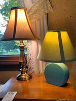 Photo of two bedside lamps on wooden surface near window with lace curtains. Left lamp has metallic base and beige fabric shade with brown trim. Right lamp has round blue ceramic base with beige fabric shade accented with dark trim.