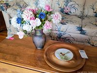 Photo of the Ruben hand painted glass vase with a floral arrangement, next to a signed wooden serving tray with a bird illustration, set on a wooden coffee table with a floral couch background.