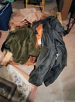 Two vintage pilot clothing items, a green jacket and a greenish-grey pilot suit, displayed on the floor with some background clutter.