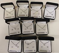 Overview of 11 black jewelry boxes each containing a silver chain necklace and matching earrings set, arranged in rows.