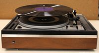 Front angled view of Dual Changer 1225 record player playing a vinyl record. Highlighting wood and metal body with platter and spindle.