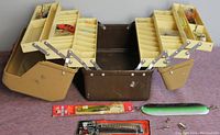 Image showing the open vintage fishing tackle box with fold-out trays and various fishing lures placed inside and around it.