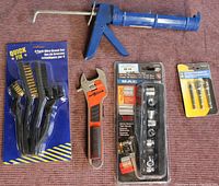 Wide shot showing manual blue caulking gun, red and black AutoWrench adjustable wrench, packaged wire brush set, socket driver adapter set, and SAE socket set.