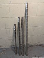 Four piano hinges of varying lengths displayed vertically against a white concrete wall. The hinges show their nickel plating and numbered length labels.