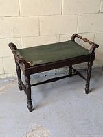 Antique wooden vanity bench with green upholstered seat, dark wood frame, spindle arm handles, and turned legs.