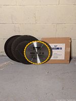 Photo showing four blades including a yellow-edged Dewalt 12 inch circular saw blade with 32 teeth, three black metal grinding blades, and a Jet-Kut cardboard box for a 12 inch saw blade.