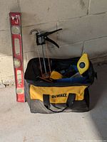 Photo showing DeWalt tool bag filled with various hand tools including a red Stanley spirit level, caulking gun, and Stanley chalk line against a concrete wall.