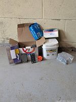 Wide photo showing the lot spread on floor with various electrical items including a cardboard box, plastic packages, and a small bucket container.