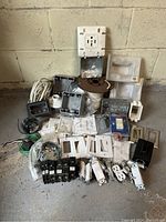Photo showing the full lot including various cover plates, junction boxes, breakers, outlets, switches, wire and more, arranged on a concrete floor against a block wall.
