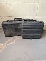 Two black hard plastic cases for laser level and cordless drill on concrete floor next to wall, cases showing wear and scuff marks