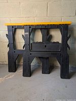 Pair of folding sawhorses shown folded and standing against a wall, yellow top rails and black legs visible.