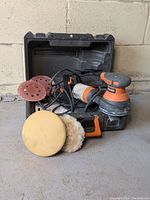 Orbital sander with power cord, sanding discs, and polishing pads arranged outside the open carrying case.