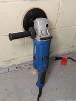 Front left angle view of the blue Simoniz polisher with black side handle and round polishing pad attachment, standing up by a concrete wall.