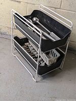 Two-tier white metal frame with black stackable plastic bins holding assorted ratchets and sockets, shown placed on concrete floor.