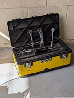 Yellow Stanley metal toolbox open with black lid, showing inside multiple hand tools including wrenches and ratchets.