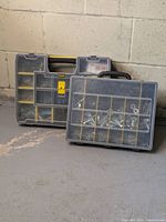 Two plastic organizer tool boxes placed on a concrete floor with a concrete wall backdrop. The Stanley box in back is 17" x 13" x 3" with a yellow latch and handle, the smaller unbranded box in front is 14" x 11" x 3" with a visible assortment of metal fasteners inside.
