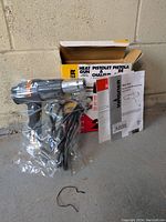 Heat gun in plastic wrap with power cord, paper manuals, and original box against concrete wall.