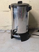 Full front view of the stainless steel cylindrical coffee urn with black handles and lid, standing on a concrete floor.