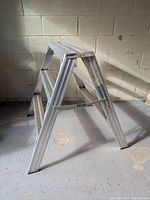 Front-angled view of the aluminum widebody step ladder showing three wide, textured steps and A-frame construction in a concrete room.