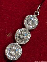 Close-up photo of the pendant showing the three round-cut Moissanite stones vertically aligned within simple bezel settings on a red fabric background.