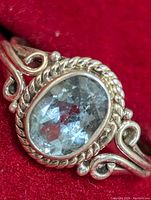 Close-up of the silver ring with an oval faceted blue topaz stone set in decorative bezel and swirl band design on red background.