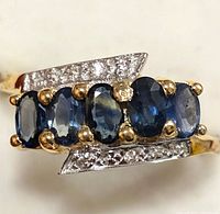 Close-up front view showing five oval blue sapphires aligned horizontally on a silver ring with small diamonds on arrowhead-shaped settings on each side.