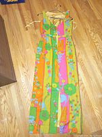 Front view of a colorful handmade sleeveless maxi dress from the 1960s with floral pattern and button front bodice, hanging on a hanger on a wooden floor.