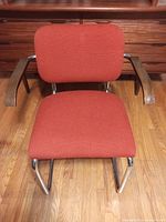 Front view of the orange upholstered office chair showing the seat, backrest, wooden armrests, and steel chrome tubular frame