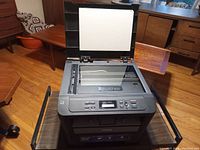 Image showing black Brother printer and scanner with the lid open revealing the scanning bed and control panel featuring buttons and a small display screen.