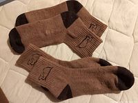 Two pairs of brown camel wool socks with darker toes and heels, ribbed cuffs, and camel logo stitched on the ankle area.